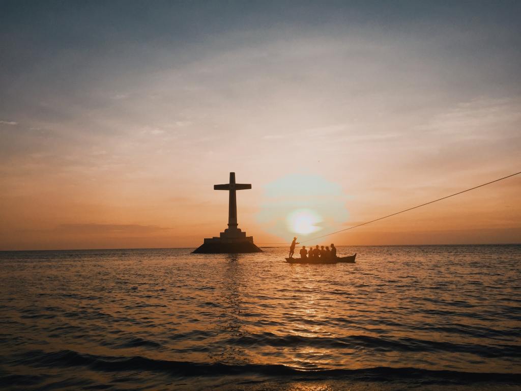 Pilgrims in small boat heading to  a big Cross in the middle of a lake at sunrise.